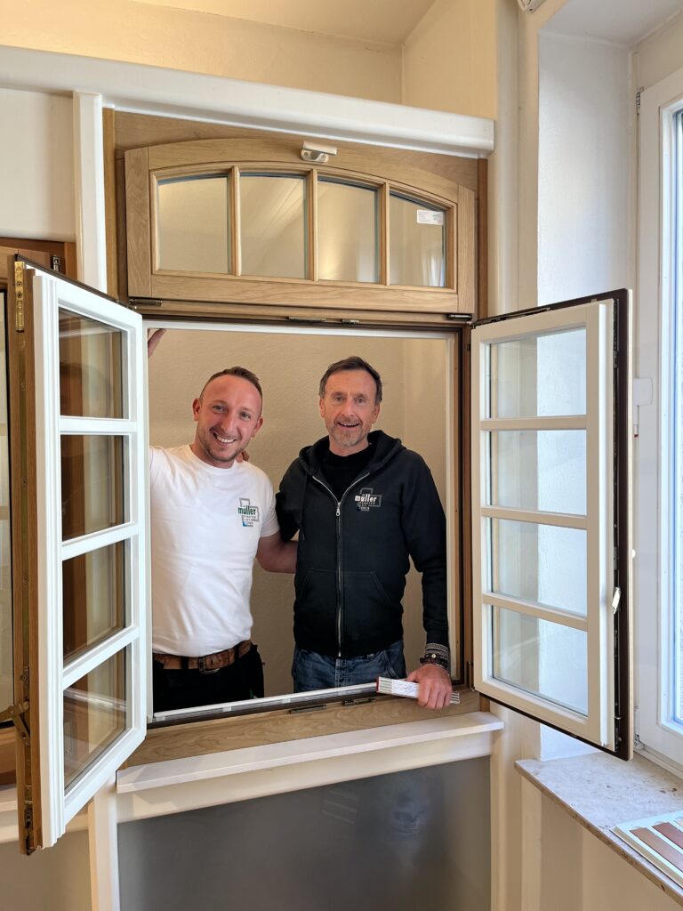 Zwei lächelnde Männer stehen freundlich nebeneinander an einem offenen Holzfenster